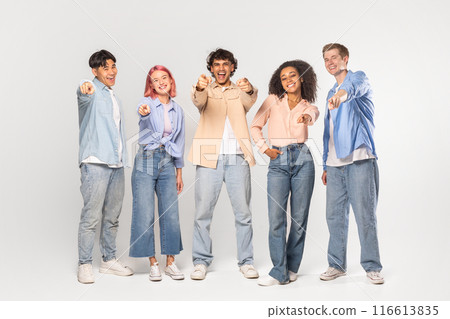 A group of five diverse friends, all smiling and pointing at the camera, are standing in front of a white background in a studio setting. 116613835