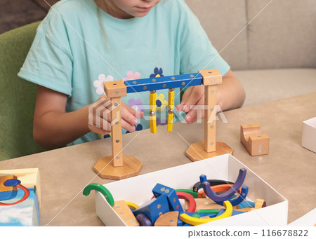 Girl assembles swing from magnetic wooden construction set. Toys and activiti children. Developmental activities at home.  116678822