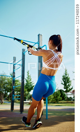 Outdoor fitness. Rear view of determined woman using TRX straps for strength training, her posture strong and focused. 117248039