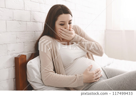 Morning sickness. Young pregnant woman sitting on bed, covering her mouth feeling nauseous during pregnancy, free space 117499433