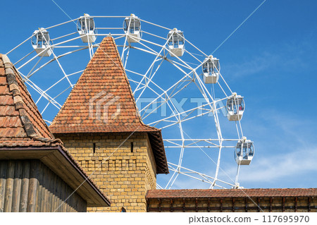 Ferris wheel towering above a historic tower on a sunny day 117695970