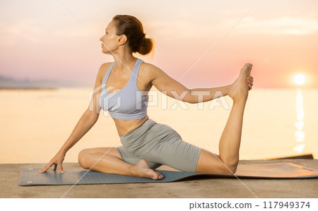 Young attractive smiling woman wearing sportswear practicing yoga, sitting in One Legged King Pigeon exercise on beach at sunset 117949374