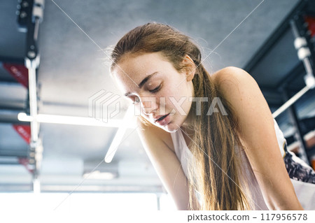 Young fitness girl with sweat on face feeling exhausted after hard workout in gym, holding knees, breathing. 117956578