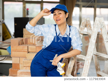 Portrait of positive builder woman in blue overalls next to stepladder 117983904