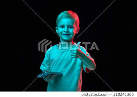 Portrait of cute little boy, child in t-shirt, holding mobile phone and raising finger up, thumbs up against black studio background in neon light 118245366