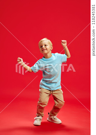 Full-length portrait of positive happy little boy, child in blue t-shirt and brown pants dancing, having fun against red studio background 118245381