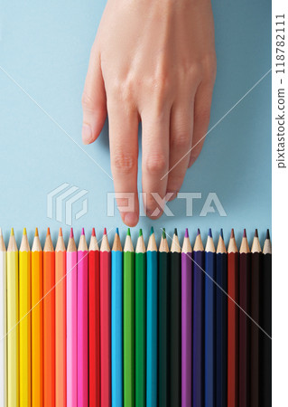 Hand choosing green colored pencil from a row of multi-colored pencils on light blue background. 118782111