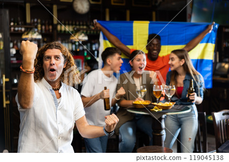 Cheerful international sport football fans holding up Swedish flag and drinking beer, eating snacks in the sport bar 119045138