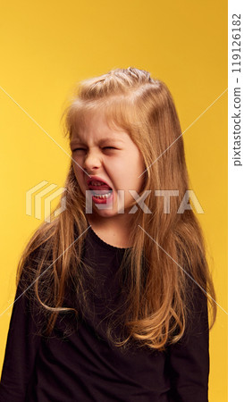 Little girl opens her mouth wide in energetic shout, her expression full of emotion and fun against yellow studio background. 119126182