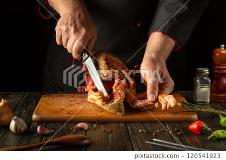 In the cozy kitchen, an experienced chef expertly slices the flavorful meat of a poultry carcass with a sharp knife. Aromatic ingredients surround the wooden table, enhancing the culinary experience 120541923