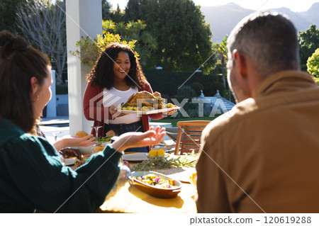 Happy diverse male and female friends serving thanksgiving celebration meal in sunny garden 120619288