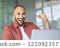 Smiling young man with joyful expression celebrates success while working on his laptop in an office environment. Concept of achievement, happiness, and professional success in a modern workspace. 121092357