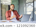 Young professional using laptop gesturing expressively during online meeting. Confused and frustrated looks, showing concern. Modern office setting indicates technology and communication challenges. 121092360