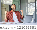 Joyful man with laptop showing excitement, celebrating success. Modern office setting emphasizes achievement and happiness. Positive vibes and energy are evident in his expression. 121092361