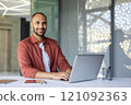 Smiling man working on laptop at desk in modern office, showcasing productivity. Table holds smartphone and pen holder, reflecting efficiency and work environment. Bright, professional setting. 121092363