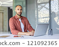 A professional man seated at a desk working on a laptop in a modern workspace, conveying focus and productivity. Includes office supplies and a bright, open environment. 121092365