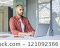 Confident businessman working diligently on a laptop in a bright, modern office. The professional setting includes essentials like a notebook, pen, and tablet, emphasizing focus and productivity. 121092366