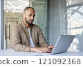 Professional man using laptop in office setting, showing concentration on work tasks. Modern workspace highlights focus, technology, and business environment. 121092368