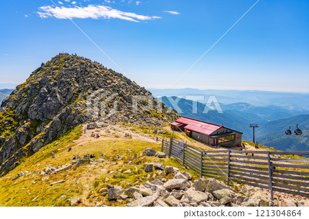 Visitors relax and enjoy breathtaking views atop Chopok Mountain in the Low Tatras, Slovakia. The sunny day highlights the rocky terrain and picturesque cabin nearby. 121304864