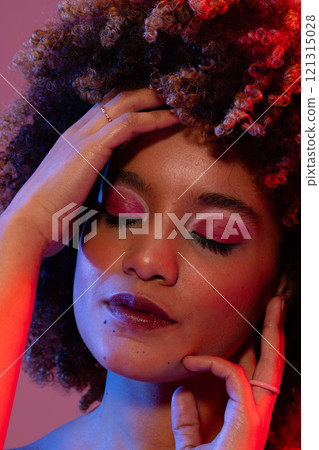 Biracial woman wearing eyeshadow and lipstick touching face with eyes closed in red and blue light 121315028