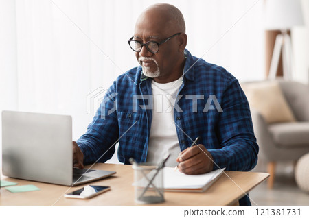 Senior Black Man Using Laptop And Writing Report Taking Notes Working Online Sitting At Desk In Office, Wearing Eyeglasses. Entrepreneurship In Mature Age, Business Lifestyle Concept 121381731