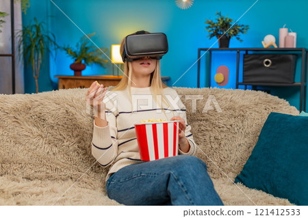 Excited woman girl wears VR headset watching movie film eating popcorn sitting at home on sofa 121412533