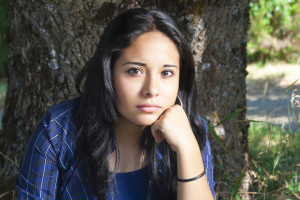 A person sitting against a tree looks concerned as they rest their head upon their fist.