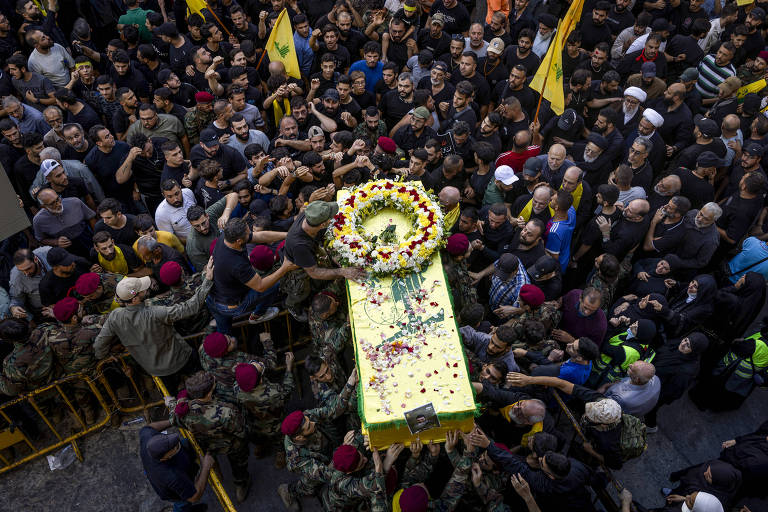 A imagem mostra uma multidão reunida em um funeral, com pessoas vestindo roupas escuras. No centro, um caixão coberto com flores e um pano amarelo é carregado por várias pessoas. Há bandeiras do Hezbollah visíveis entre a multidão. A cena é marcada por expressões de luto e solidariedade, com muitos homens e mulheres ao redor do caixão, alguns em uniformes militares.