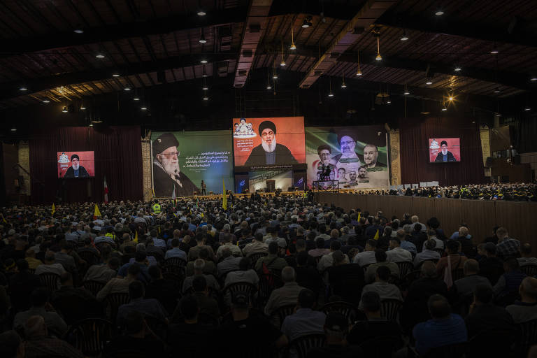 A imagem mostra uma grande multidão reunida em um evento, com várias telas exibindo discursos. No fundo, há painéis com imagens de líderes e figuras importantes, incluindo um homem de barba e turbante. A audiência é composta principalmente por homens, muitos dos quais estão usando camisas de cores variadas. Bandeiras e símbolos estão visíveis entre os participantes, e a iluminação do local é suave.