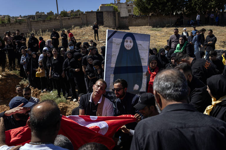 A imagem mostra uma cerimônia de luto ao ar livre, com um grupo de pessoas vestindo roupas pretas. No centro, dois homens seguram uma bandeira, enquanto um deles parece estar em um estado emocional intenso. Ao fundo, uma mulher segura um retrato de uma menina, que está vestida com um hijab e uma faixa preta. O ambiente é um terreno árido, com um muro ao fundo e várias pessoas observando a cena.