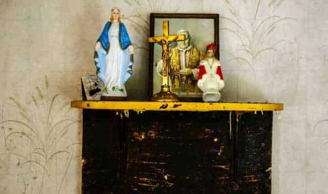 An altar shelf featuring religious items, including a cross, statues of the Virgin Mary and another figure, and a framed picture, against a wall with subtle decorative patterns.