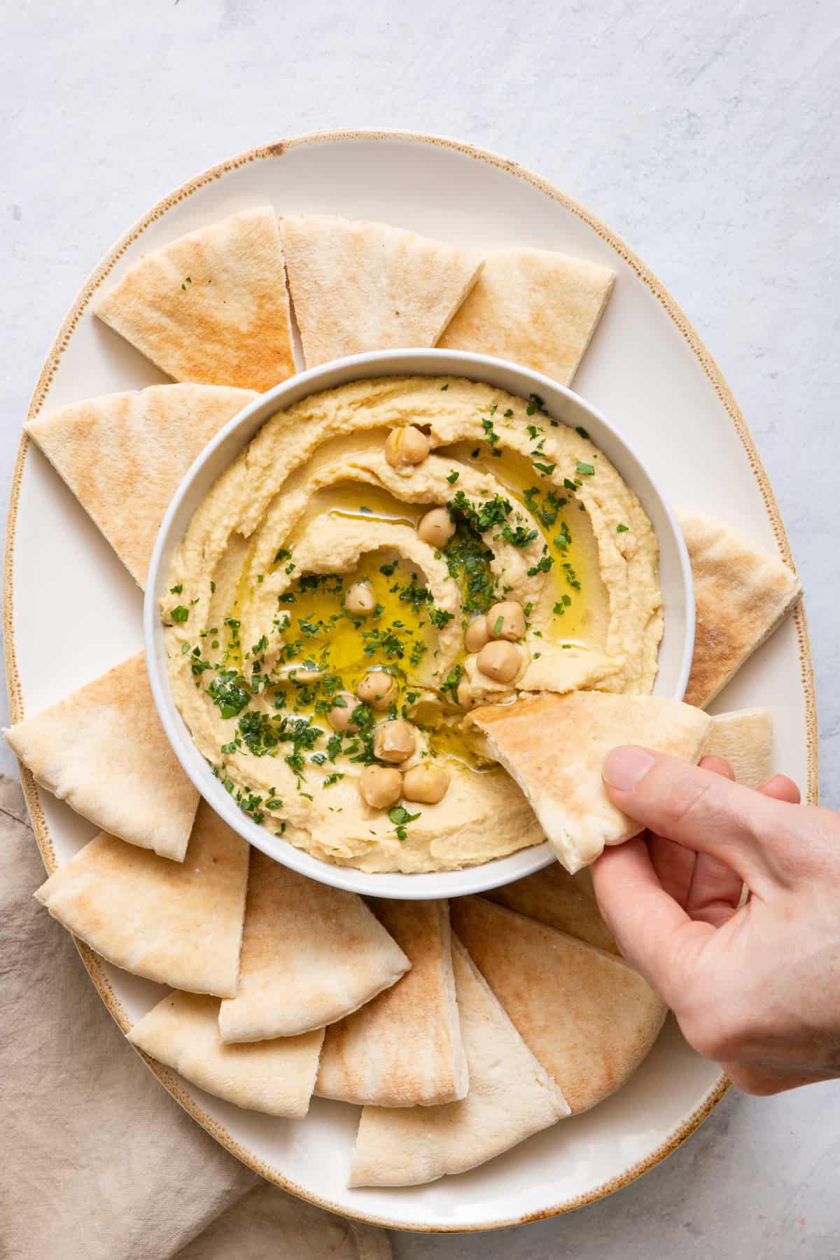 Serving plate with bowl of hummus with pita chips around bowl and a hand using a pita to scoop out a serving.