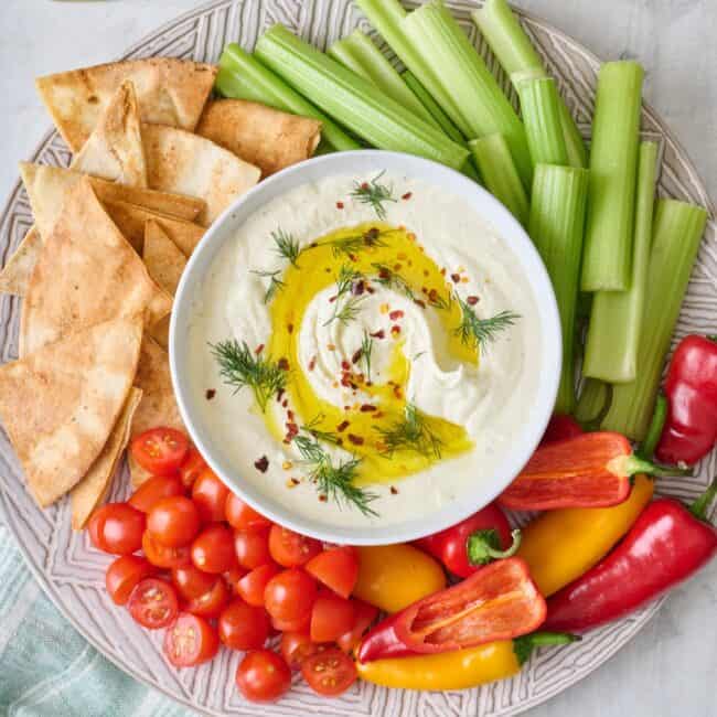 Platter of pita chips and fresh veggies with bowl of feta dip in center.