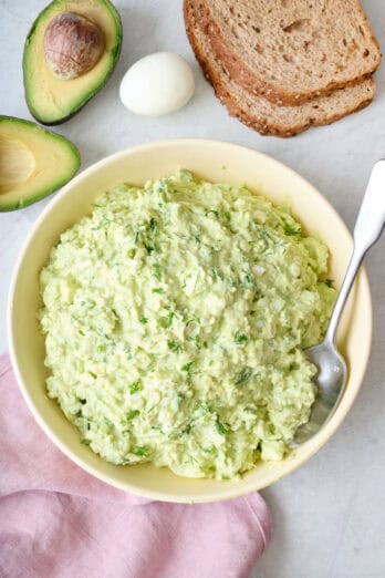 Avocado egg salad on a bowl with spoon dipped inside, fresh avocado, a boiled egg, and bread slices nearby.