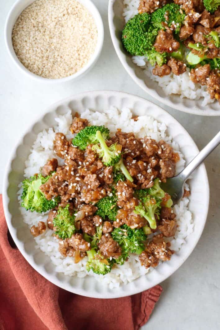 Two shallow bowls of beef and broccoli served centered over rice with fork and small dish of sesame seeds nearby.