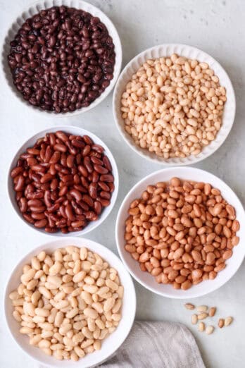 Five different types of cooked beans in separate bowls: black beans, navy beans, kidney beans, pinto beans, and cannellini beans.