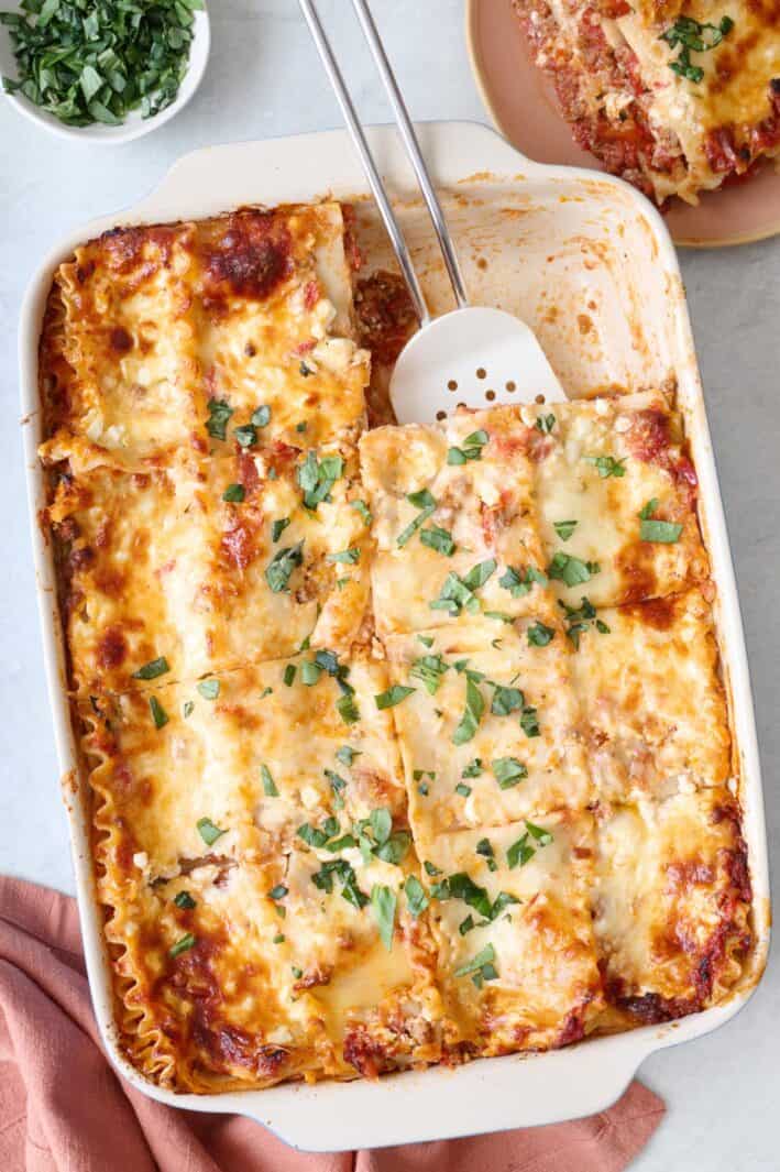 Lasagna made with cottage cheese and meat sauce in a baking dish after cutting with a large serving removed to a plate nearby.