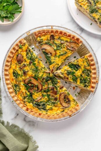 Quiche in pie plate with 2-3 slices cut, garnished with extra fresh herbs. One slice removed to a plate nearby.