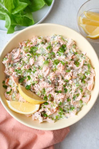 Salmon salad in a shallow serving bowl garnished with extra fresh dill and green onions, lemon slices and a plate with lettuce cups nearby.