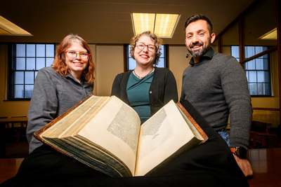 Photo of Cait Coker, Lynne M. Thomas and Elias Petrou standing in front of an open book with Greek text.