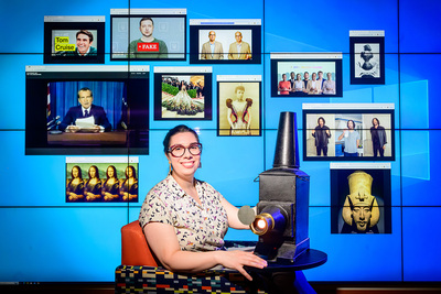 Photo of Mary Ton sitting beside a magic lantern, with a screen of deepfaked images behind her.