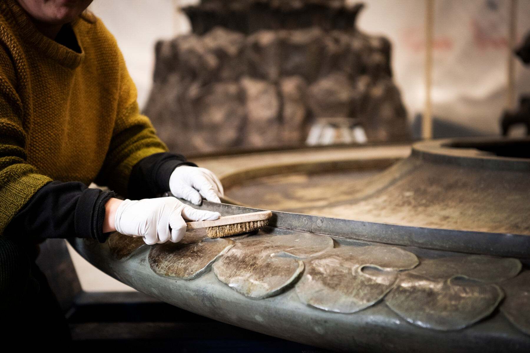 A close-up of gloved hands using a brush to clean the leaf-like patterns around one piece of the base of a metal sculpture.