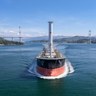 A cargo ship with an unusual cylindrical mast is sailing directly towards the camera, with islands, a bridge and mountains in the background.