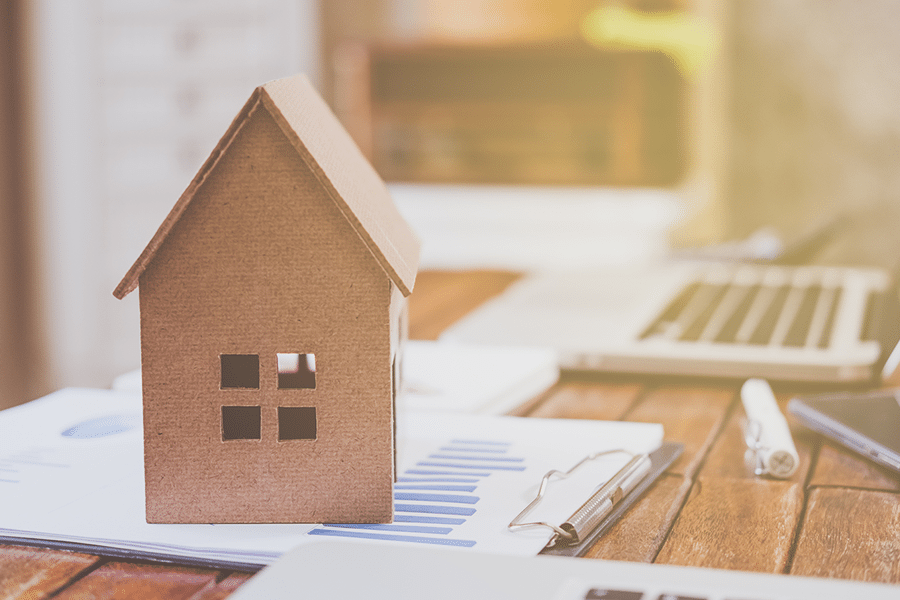 Miniature wooden house, laptop and paperwork on a table.