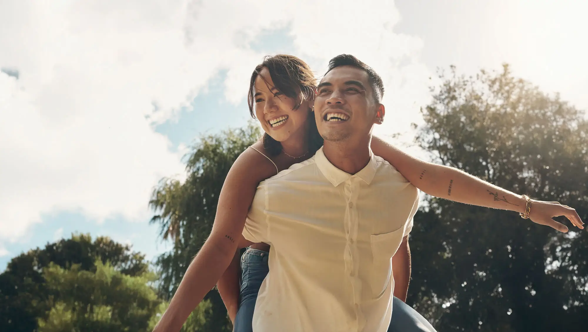 Smiling couple enjoying a playful piggyback ride in a park, surrounded by trees and a bright, clear sky.