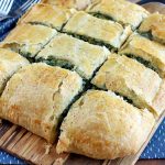 Twelve slices of spinach in puff pastry arranged in rows on a wood serving board and shot at an oblique angle, with forks on top of a blue cloth with white flecks, on a wood background.