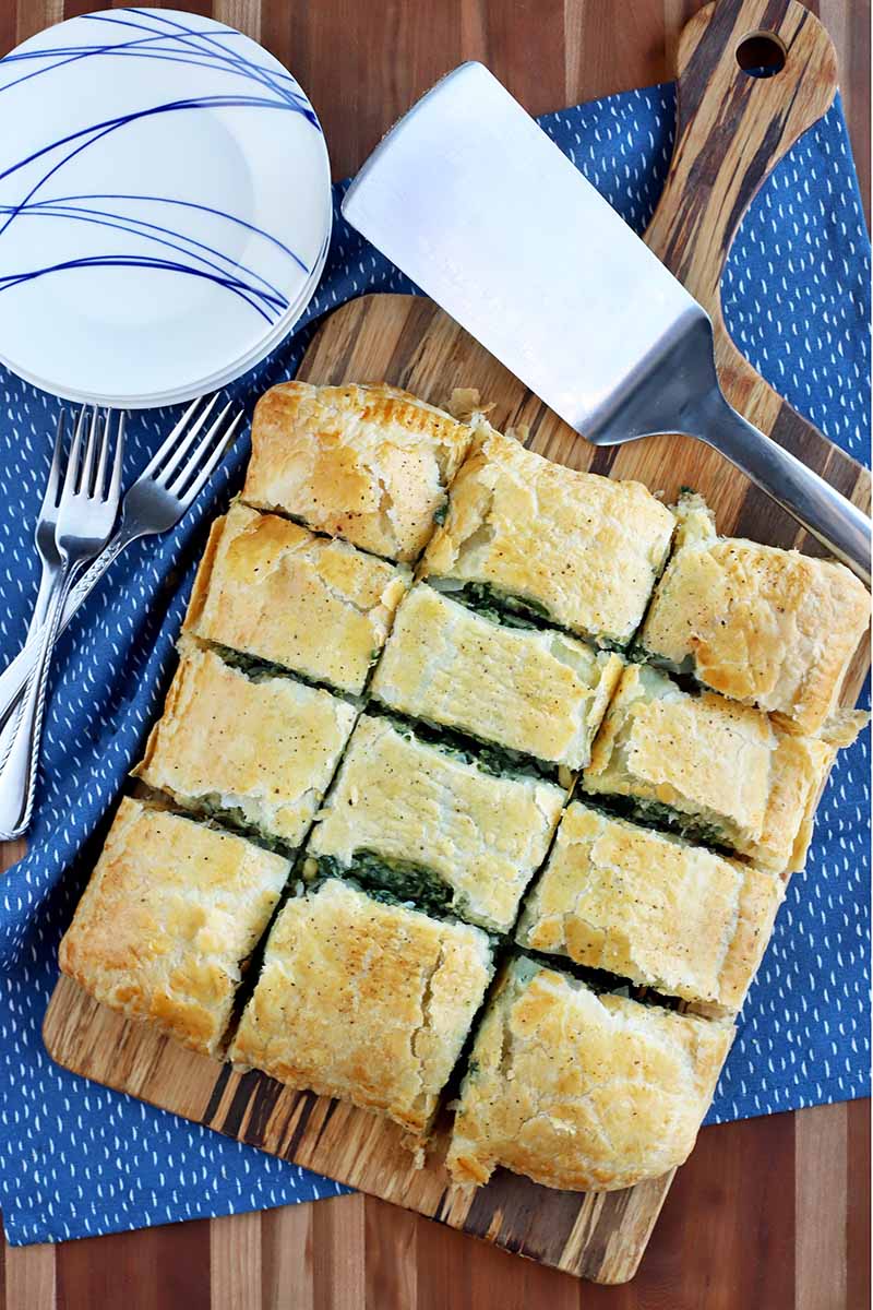 Overhead shot of homemade spinach in puff pastry cut into twelve slices and arranged on a wood board with a handle, with a metal spatula, a small stack of blue and white plates, and several forks, on a blue place mat on a striped wood table top.