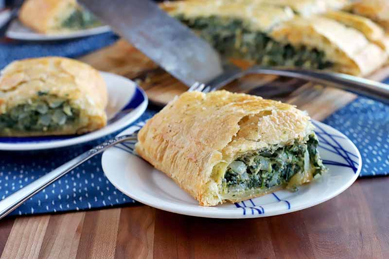 A plate of spinach in puff pastry in the foreground with more on two plates and a wood board in the background in soft focus, with a metal spatula and a blue and white place mat, on a brown wood surface.