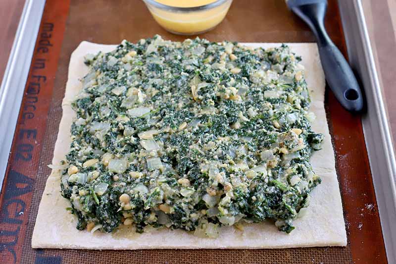 A spinach and onion mixture spread over the surface of a square piece of puff pastry with a slight border left around the perimeter, on a silicone pan liner set into a rimmed metal baking sheet, with a small glass bowl of egg wash and a black-handled silicone brush.