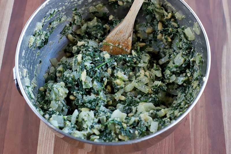 Overhead closely cropped shot of a mixture of defrosted frozen spinach, garlic, onion, pine nuts, and egg in a stainless steel bowl being mixed with a wooden spoon, on a striped brown and beige wood surface.